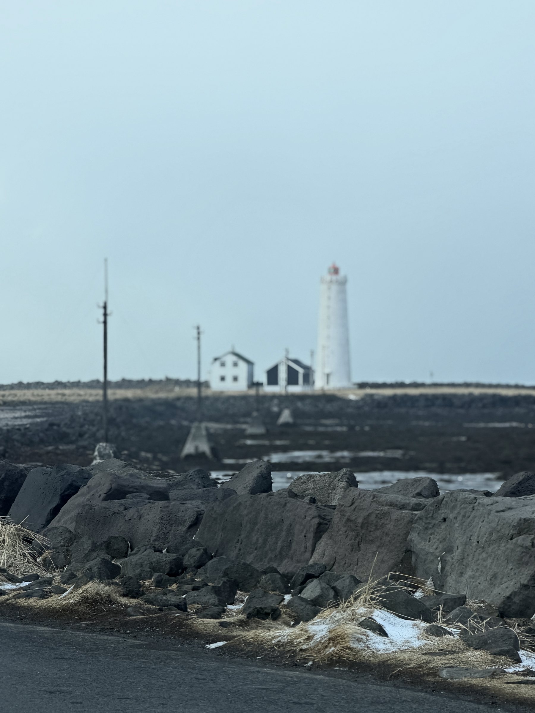 Grótta lighthouse, white against a pale sky, beyond a line of dark sea-wall rocks.