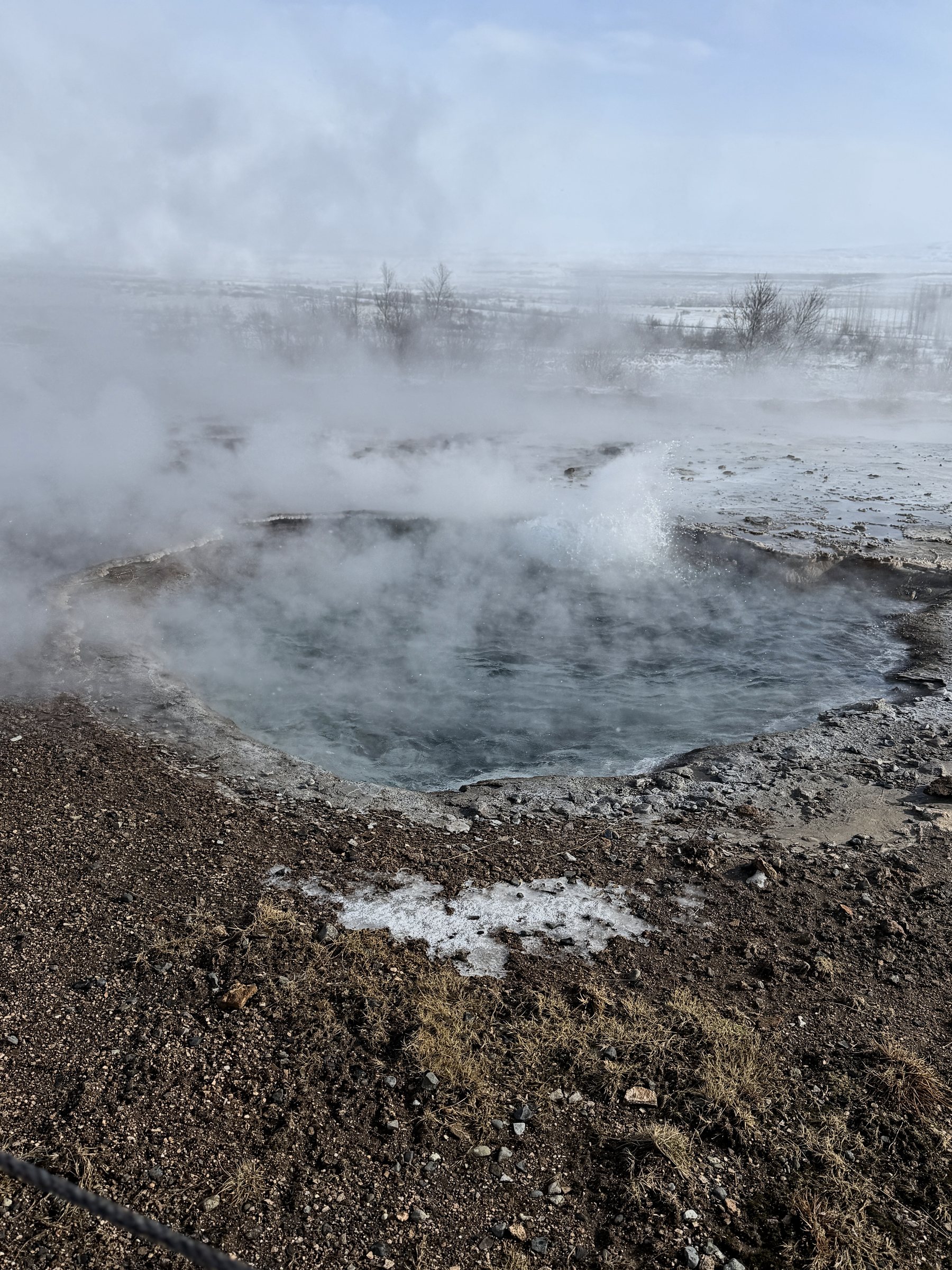 Steam rising off a hot spring pool ringed by red-brown earth and snow.