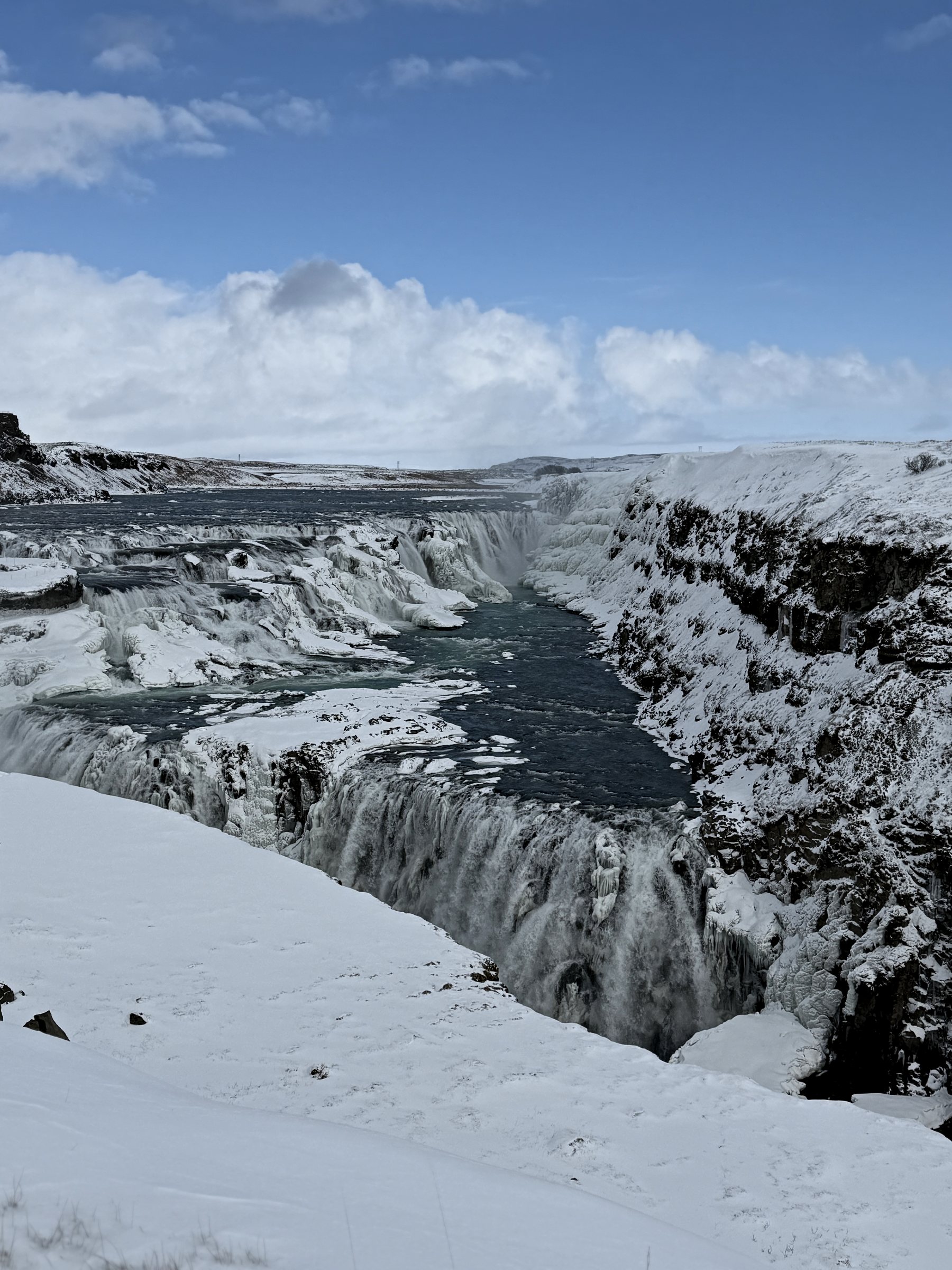 Gullfoss in winter, the falls half-frozen as they drop into a snowy canyon under blue sky.
