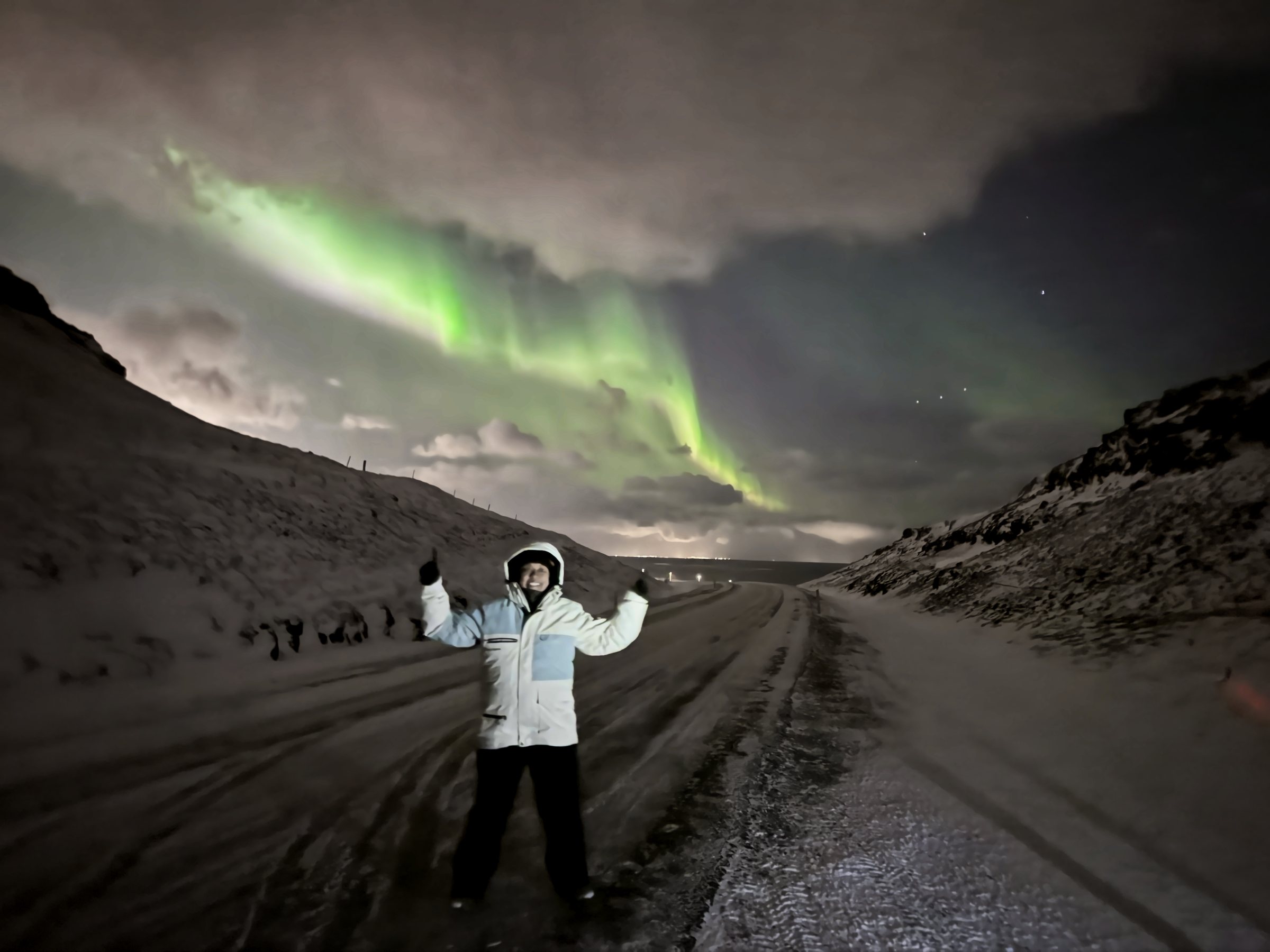 A person in a white parka with arms raised on a snowy road, a bright green aurora band crossing the sky behind.