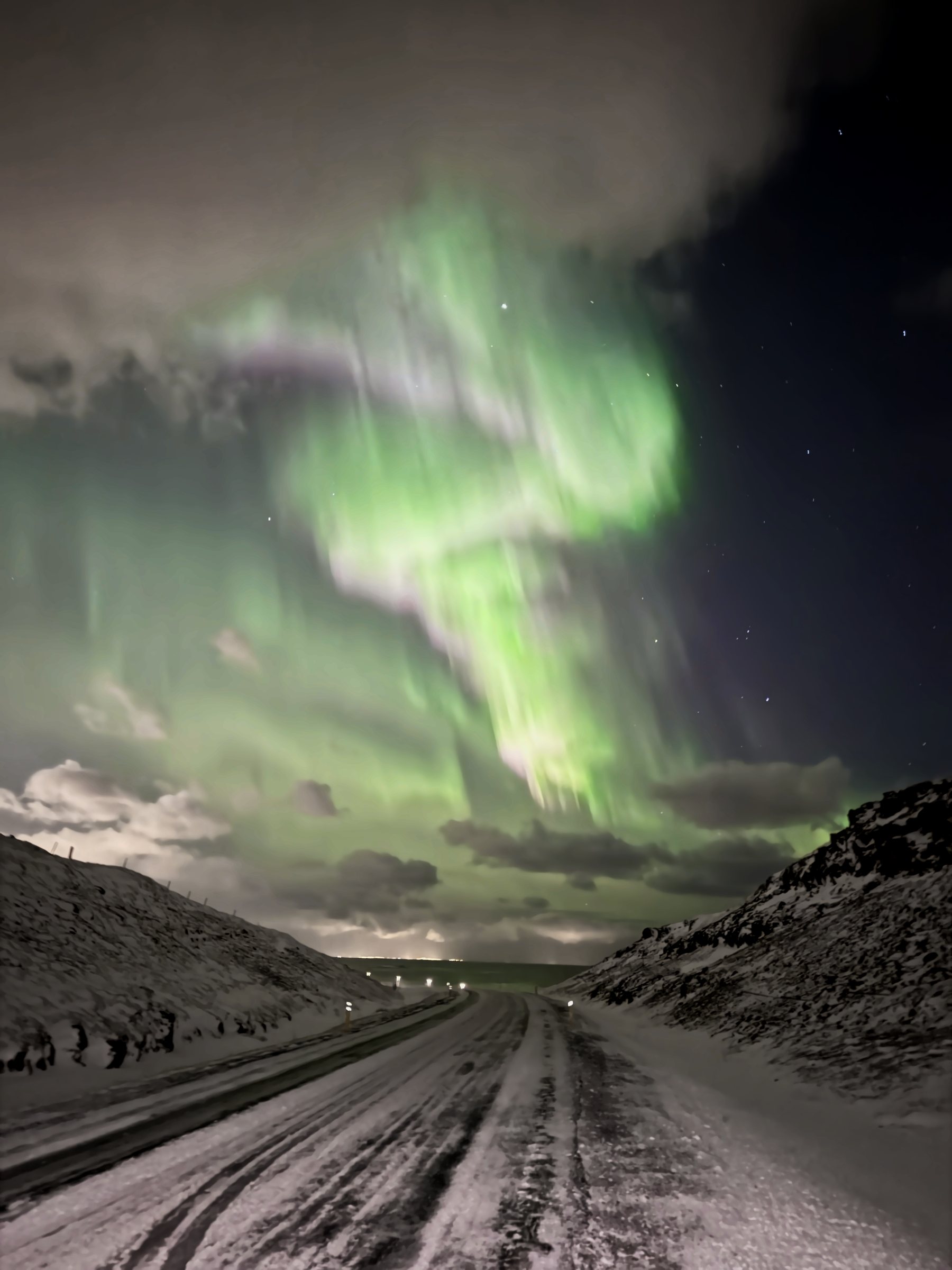 Vertical frame: aurora curtains pouring straight down from overhead toward a snowy road between hills.
