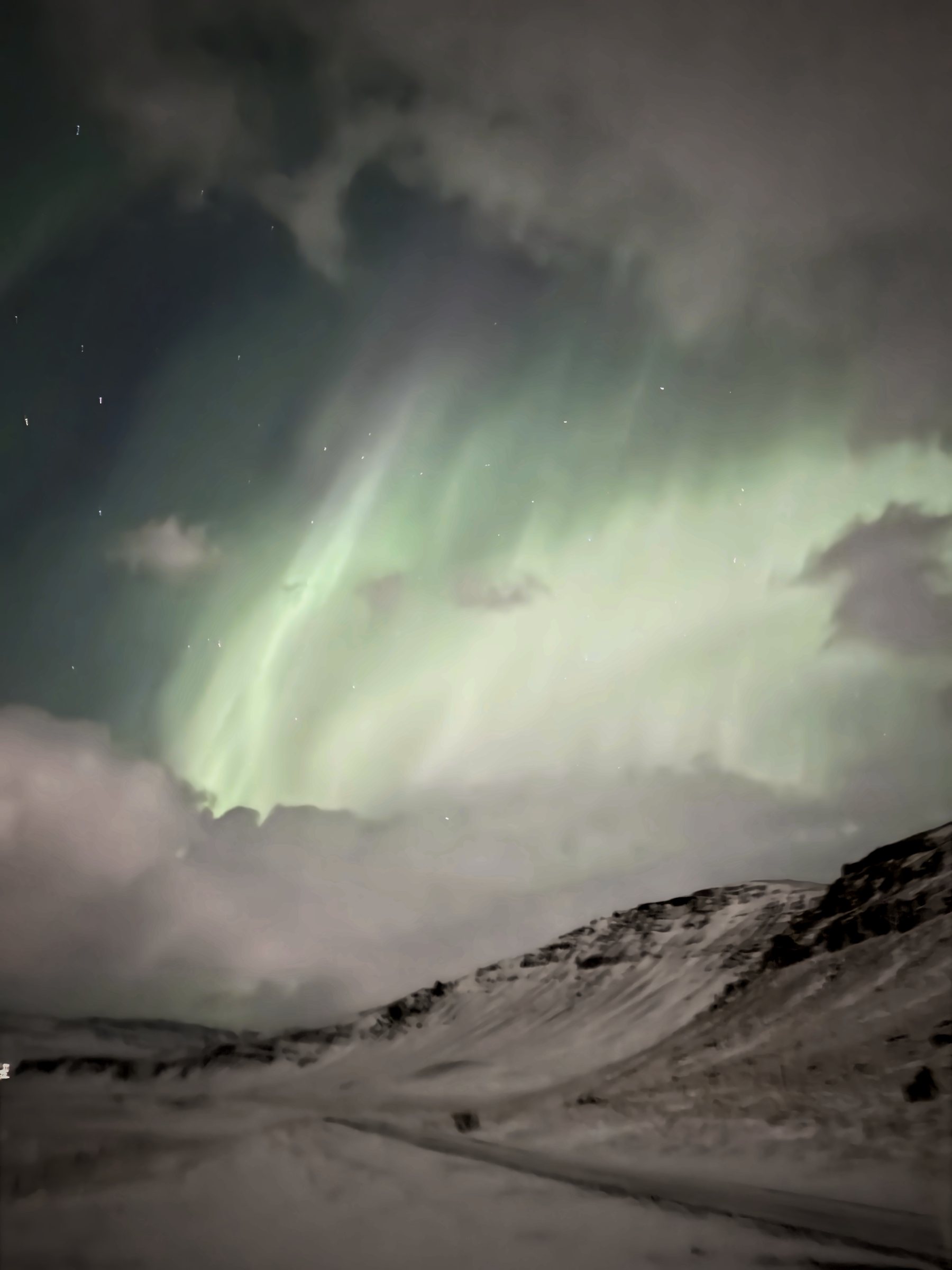 Vertical frame: a broad pale-green aurora arc breaking through cloud above a snowy hillside.