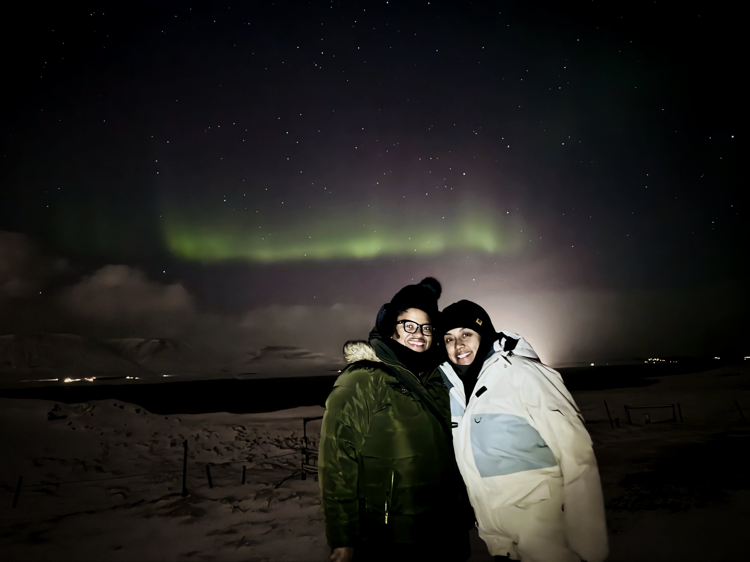 Two people in winter coats smiling under a faint green aurora arc low over dark water.