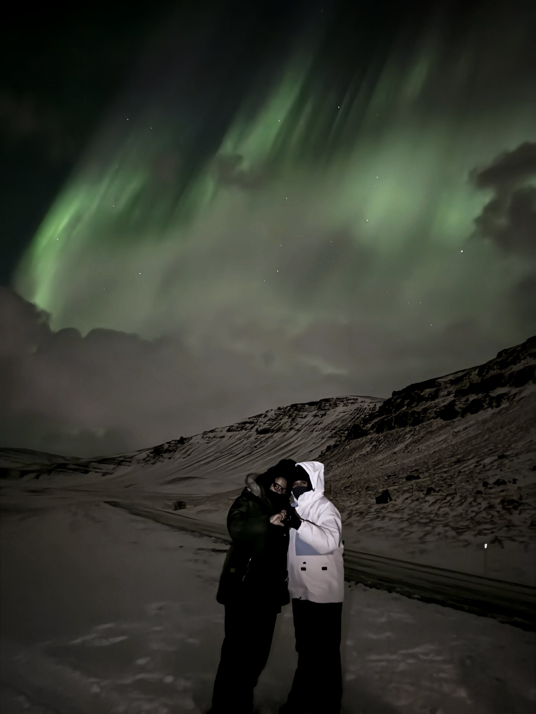 Two figures huddled together on a dark road under sweeping green aurora curtains.