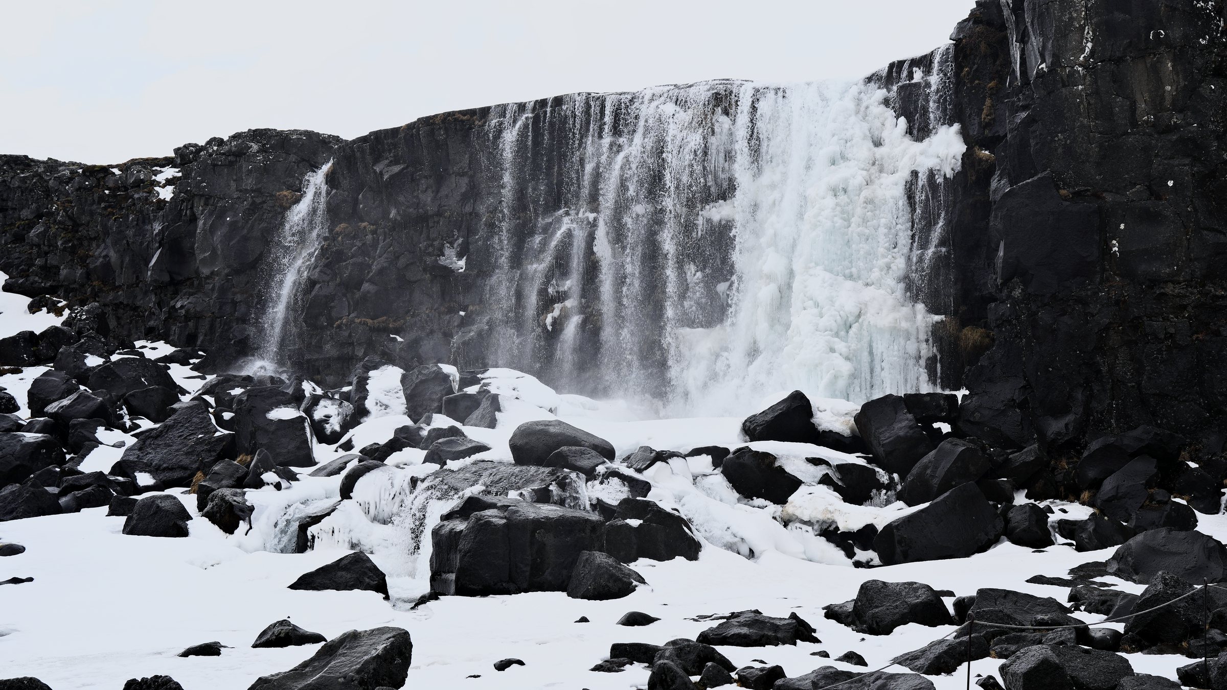 Wide view of the frozen waterfall with snow and scattered black boulders in the foreground.