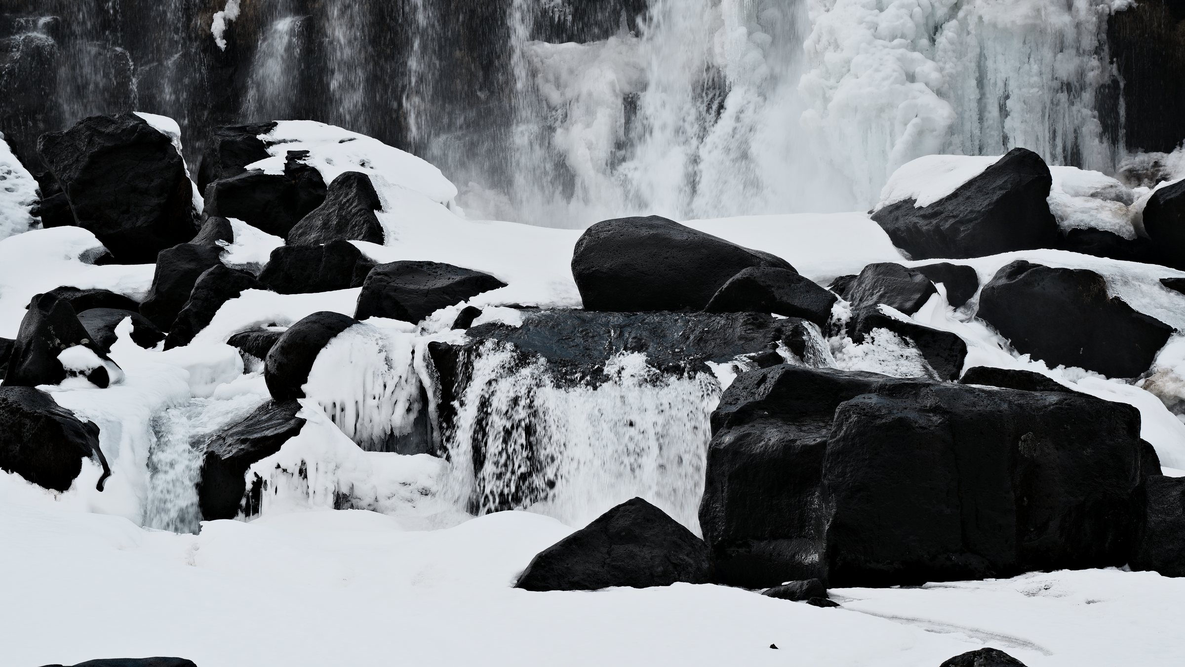 Close view of water spilling over a dark rock ledge between snow-capped boulders, edged with icicles.