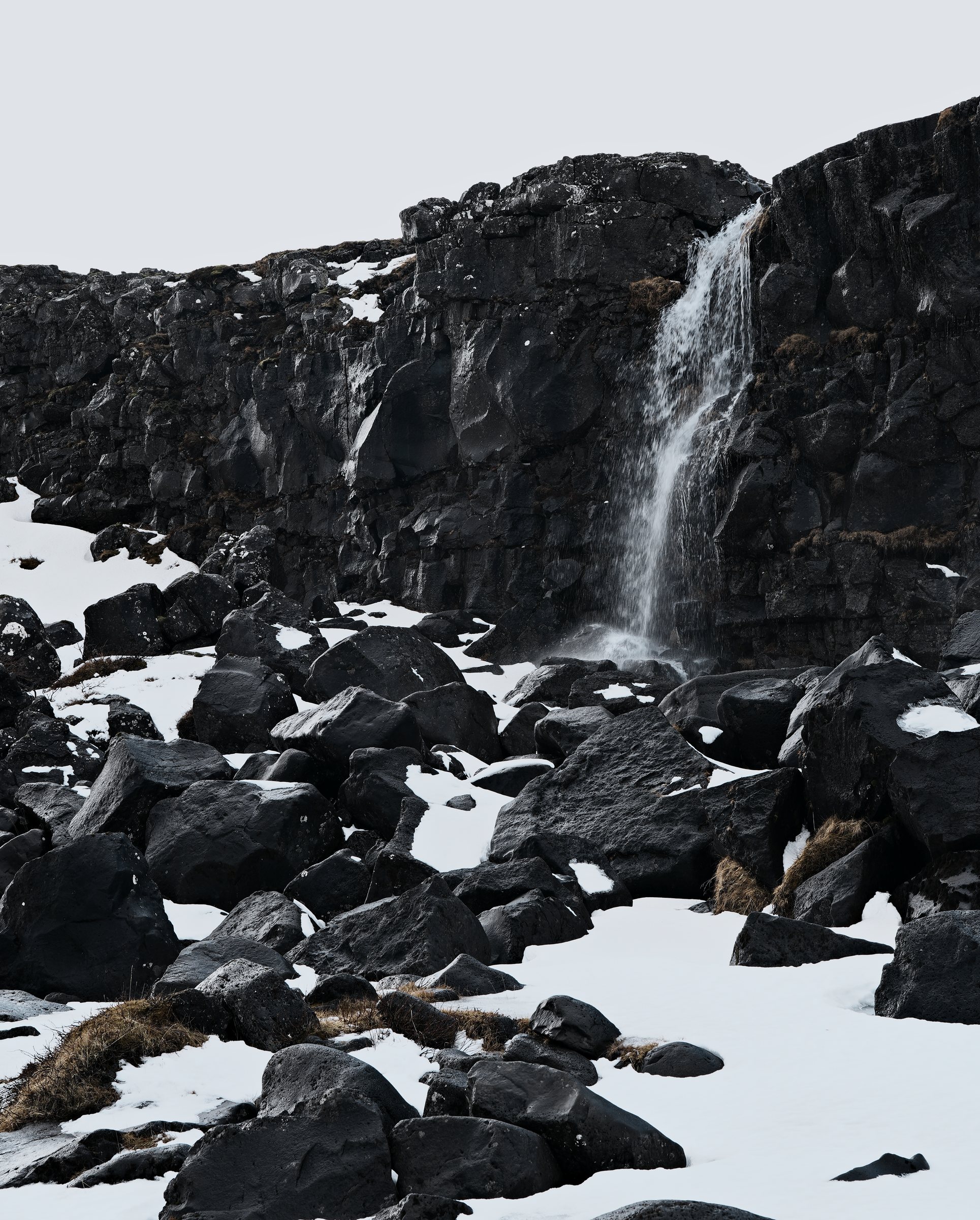 Vertical frame of a thin side stream falling down the gorge wall above a field of snow and boulders.