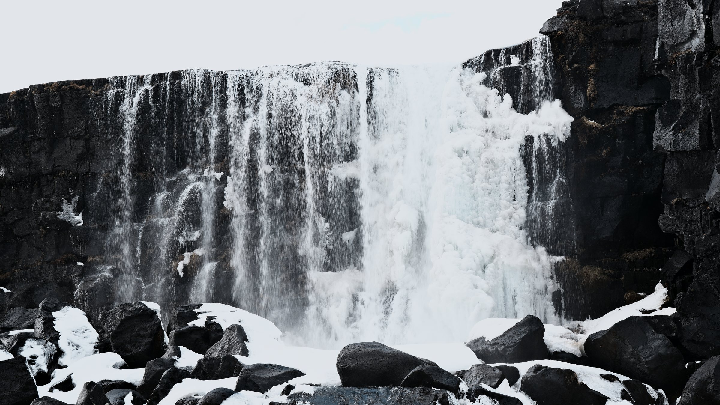 Öxarárfoss in winter: dark basalt cliffs with the waterfall frozen into columns of pale blue ice, snow-covered boulders below.