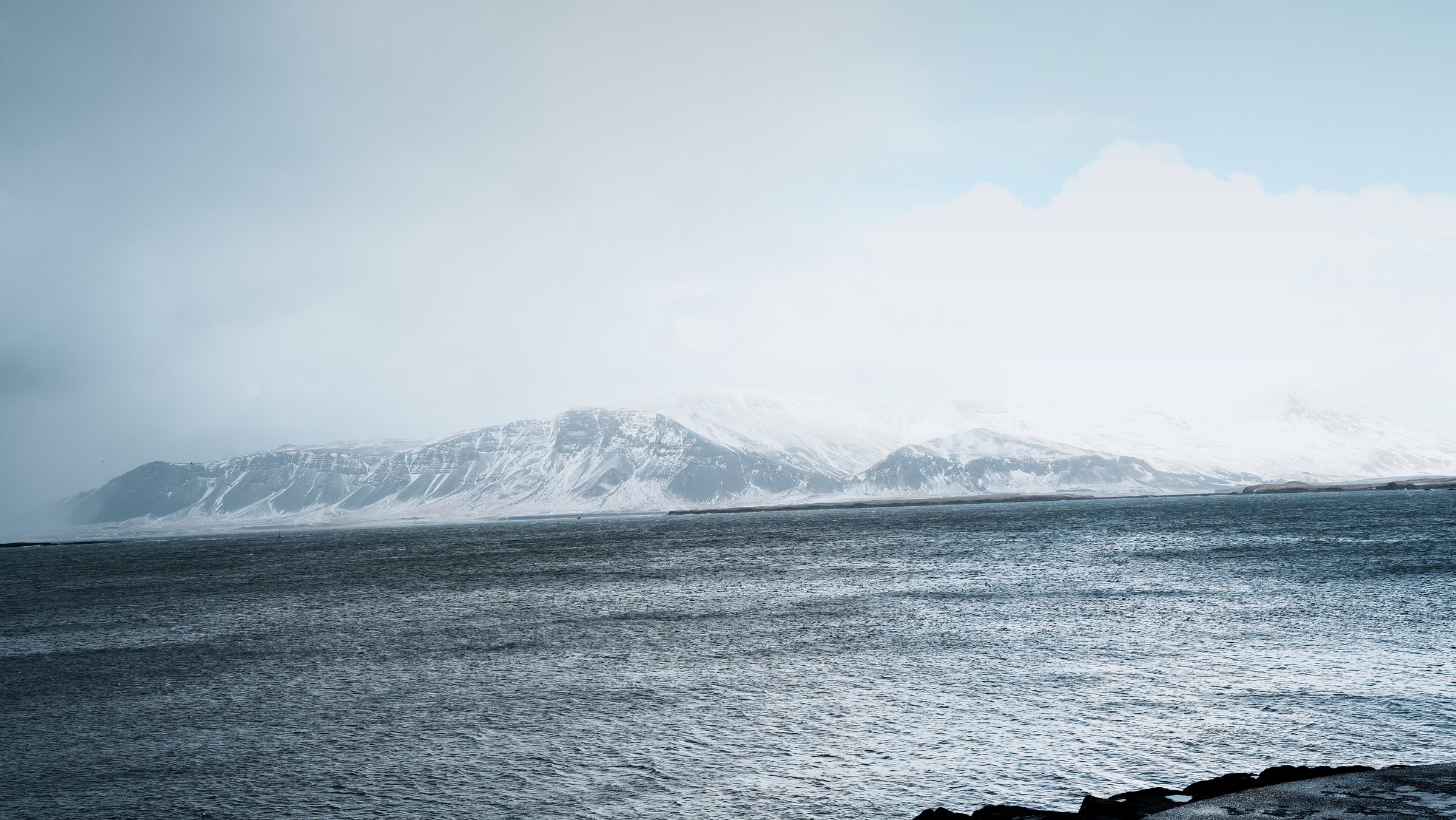 Grey-blue bay water in the foreground, a snow-covered mountain range on the far shore under heavy cloud.