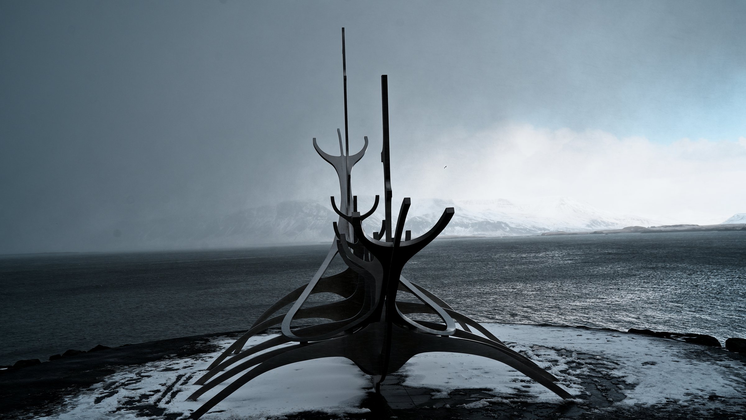 The Sun Voyager in silhouette against a dark snow squall, a band of bright light breaking over the mountains to the right.