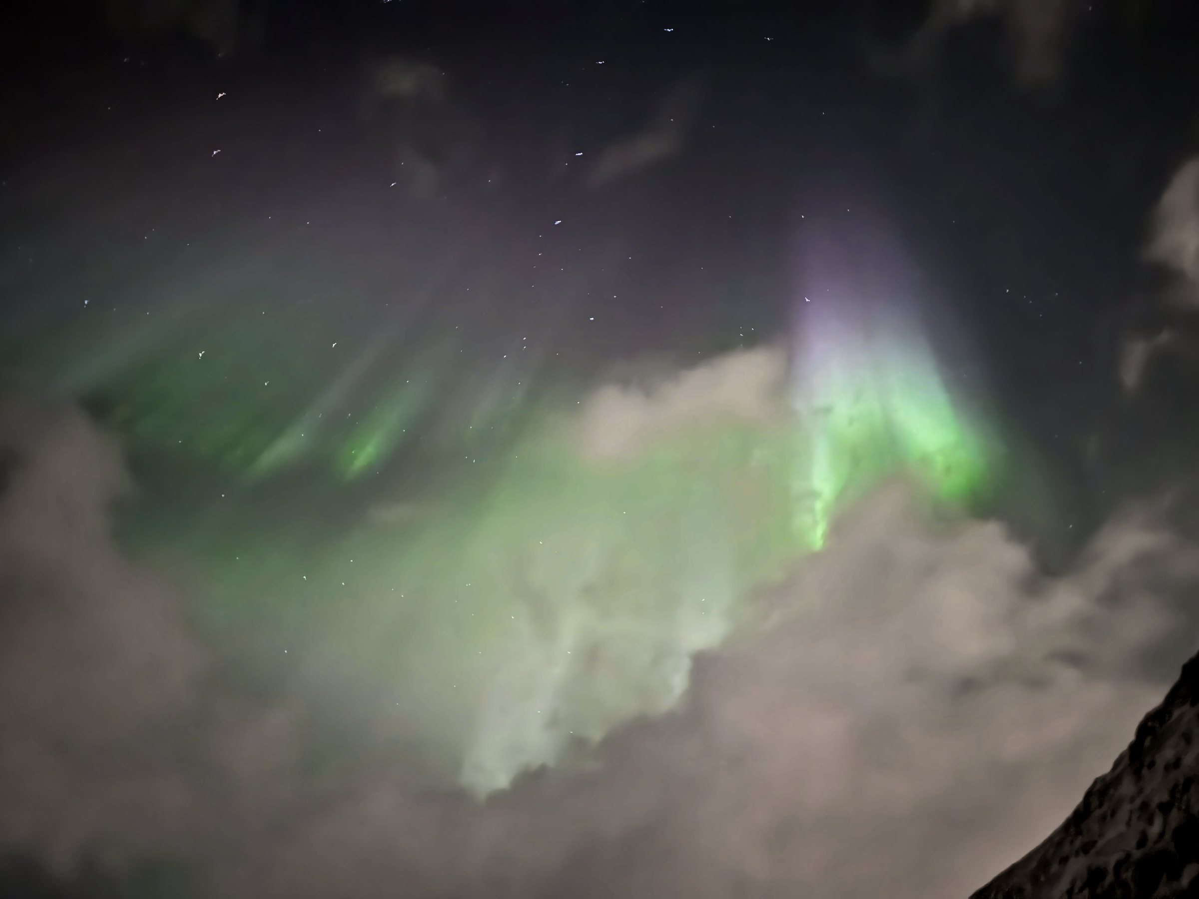 Green and violet aurora curtains hanging down behind lit clouds, stars between them.