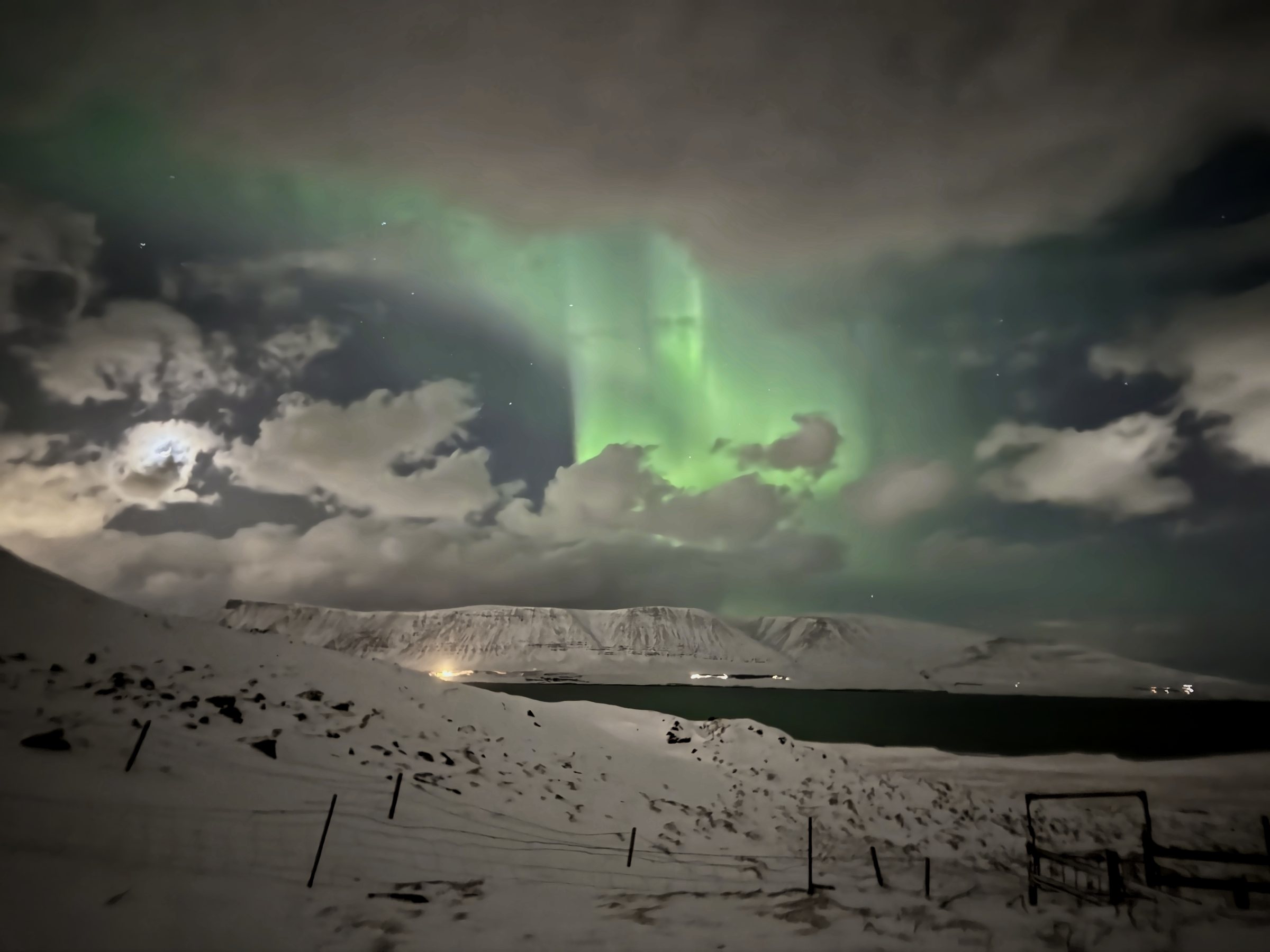 An aurora arc over dark water and snowy hills, the moon glowing through clouds to the left.