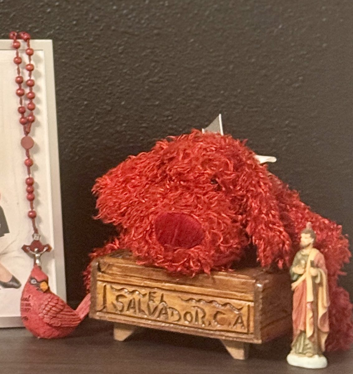 Detail of the altar: red rosary beads ending in a cardinal figurine, a carved wooden box reading El Salvador C.A., a red plush dog and a small saint statue.