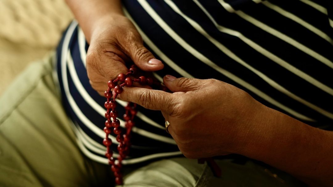 Close-up of weathered hands passing red wooden rosary beads against a navy striped shirt.