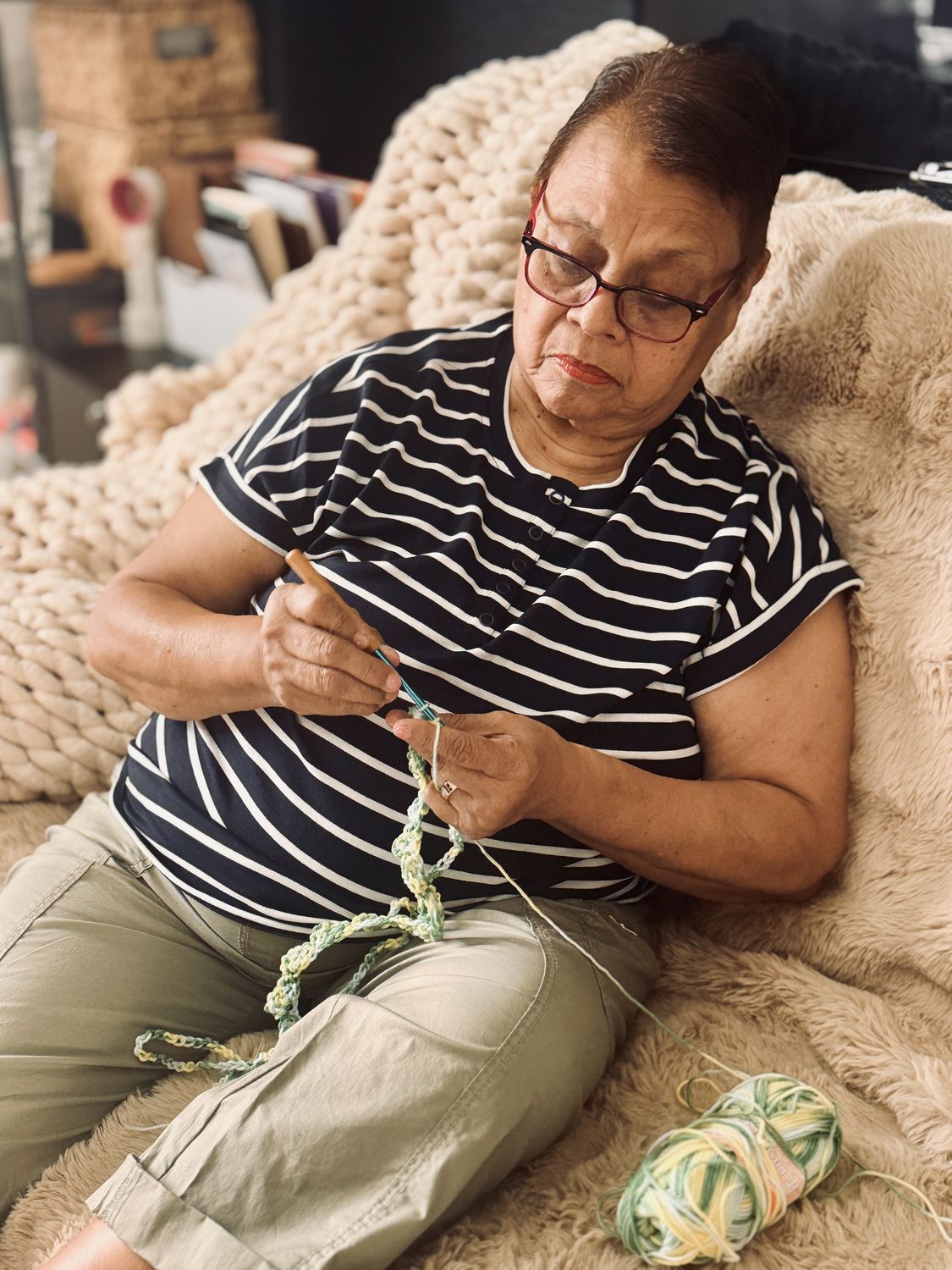 A woman in red glasses and a navy striped shirt crochets a green and yellow chain on a cream sofa, a ball of yarn beside her.