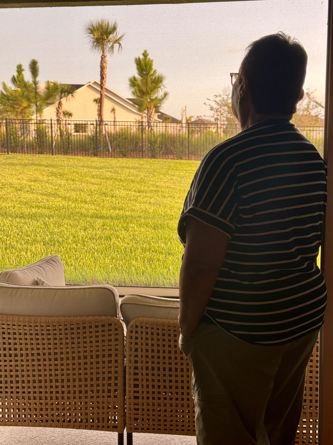 Seen from behind in silhouette, a woman stands at a screened patio door looking out at a bright green lawn, a fence, palms and a neighboring house in evening light.