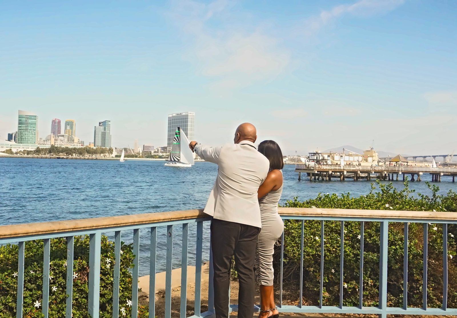 Couple from behind, he points across the bay at a sailboat