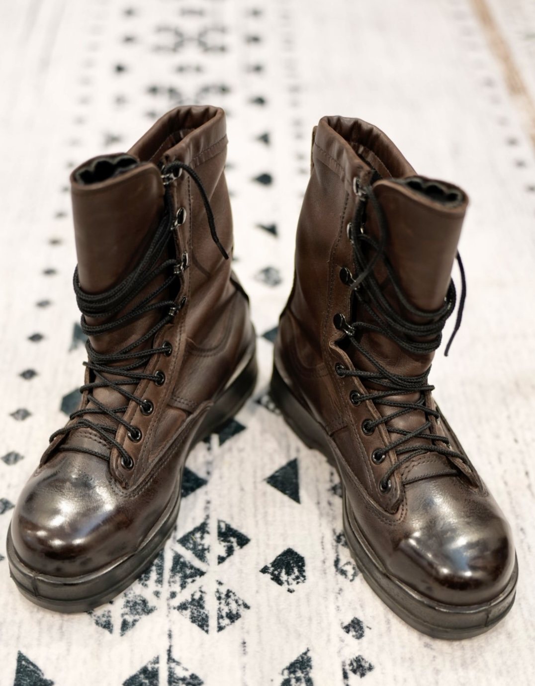 Overhead view of a pair of brown leather service boots, laces wrapped around the shafts and toe caps polished to a mirror shine, standing on a white rug with black geometric print.