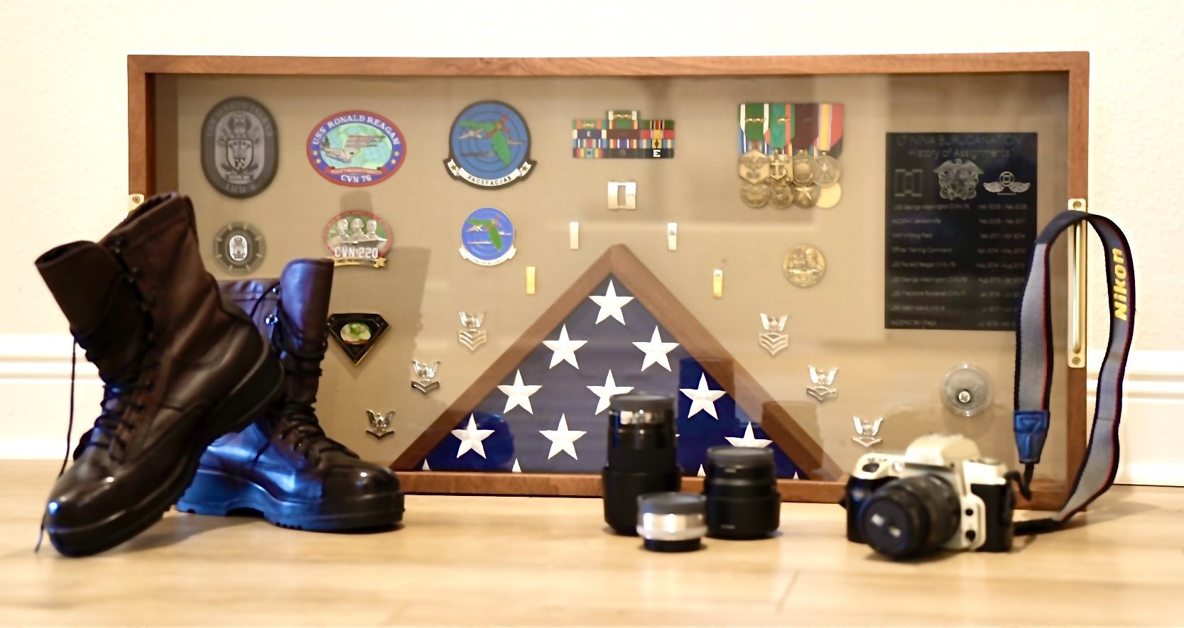 Still life on a wood floor: a walnut shadow box holding a folded flag, ship and squadron patches including USS Ronald Reagan, medals, ribbons, enlisted rating badges, officer insignia and a History of Assignments plaque; the boots stand at left, a Nikon film camera and lenses at right.