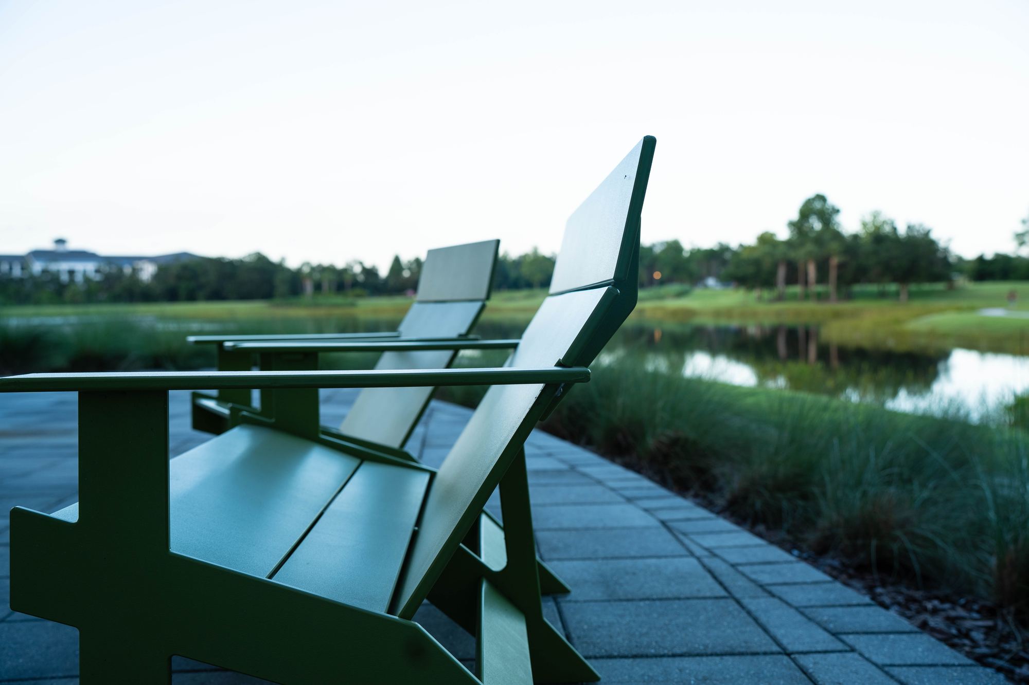 Two green modern Adirondack-style chairs on a paver walk, facing a calm lake at dusk.