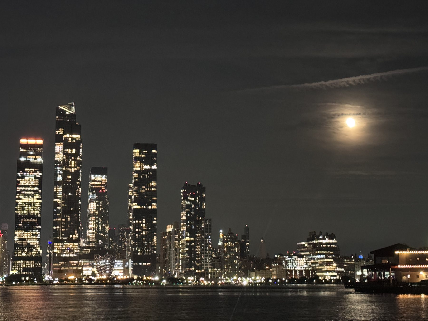 Full moon glowing through thin cloud above the lit towers of Hudson Yards, reflected in the Hudson River at night