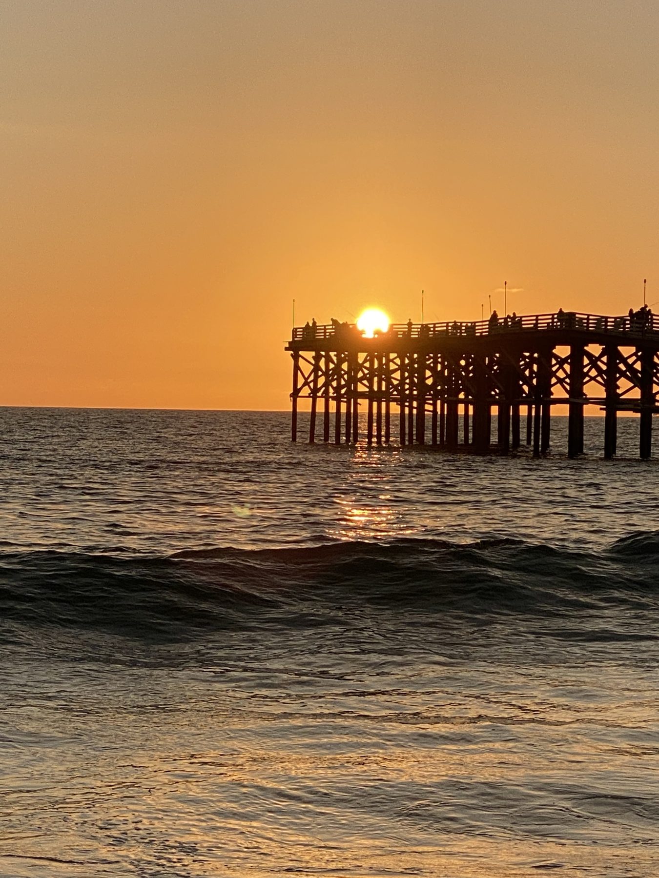 Sun setting behind the cross-braced pilings of a pier, the structure in full silhouette against an orange sky over rippled water