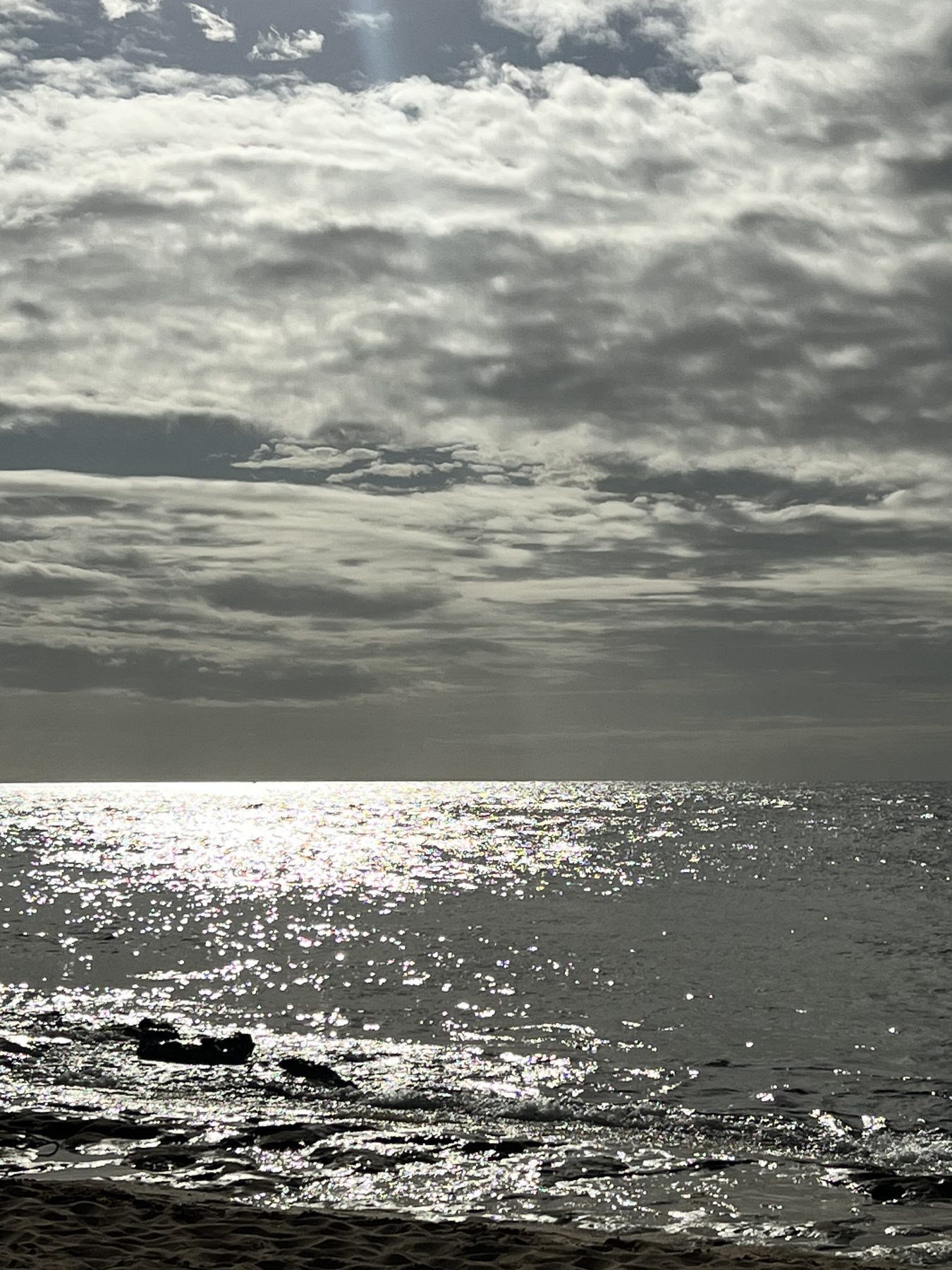 Late-afternoon sun behind heavy cloud, scattering thousands of bright points across a grey sea above a dark sandy shore